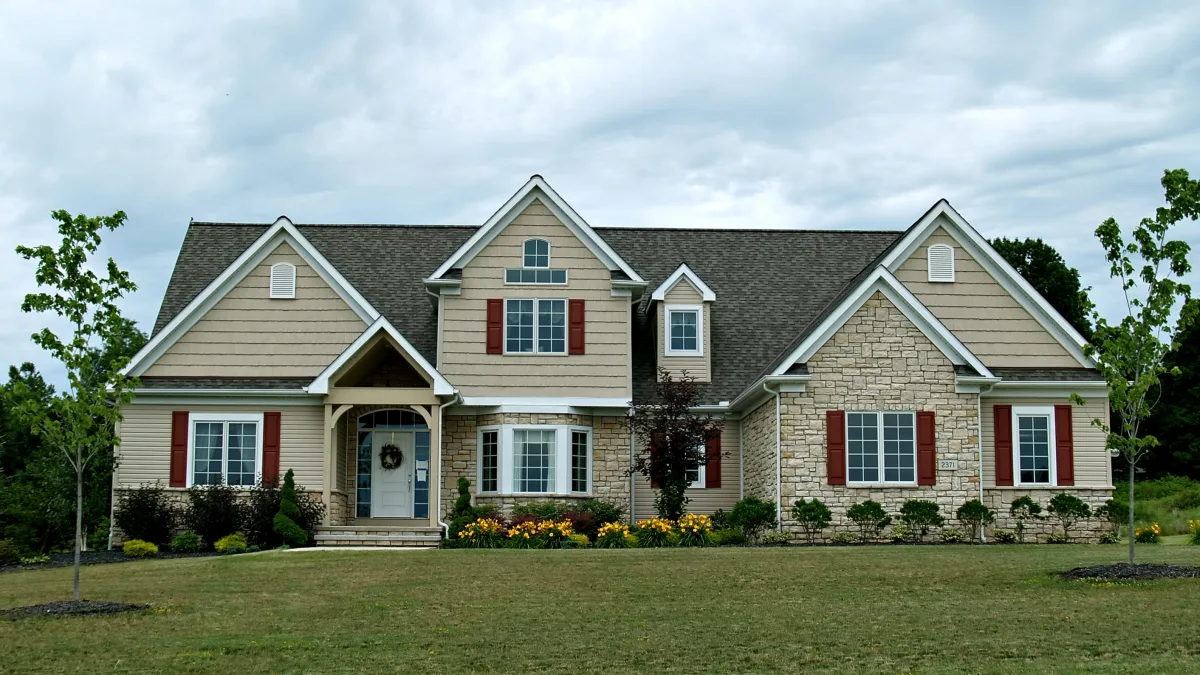 A large, two-story house with a mix of stone and siding, surrounded by a lawn and small trees during Adobestock 3643206.
