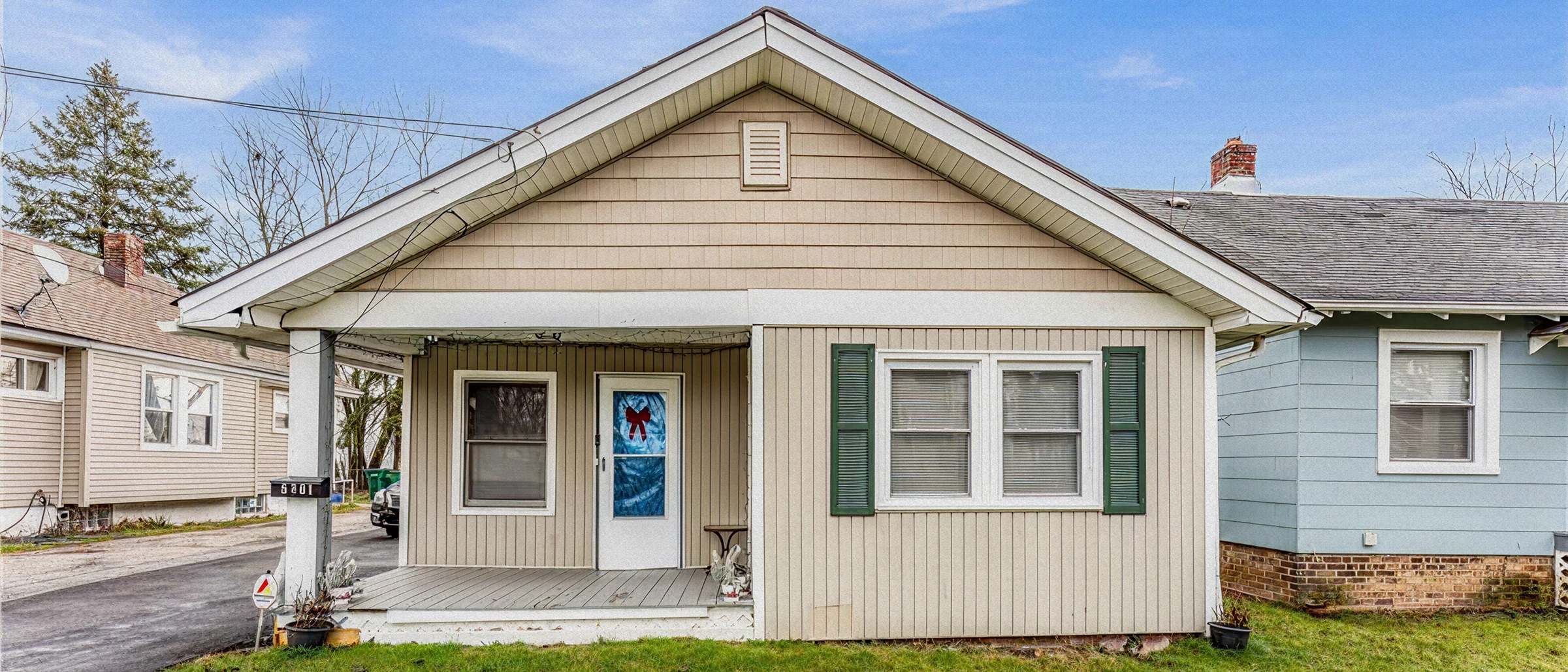 Full-width front view of a single-story house with tan siding, green shutters, and a covered front porch