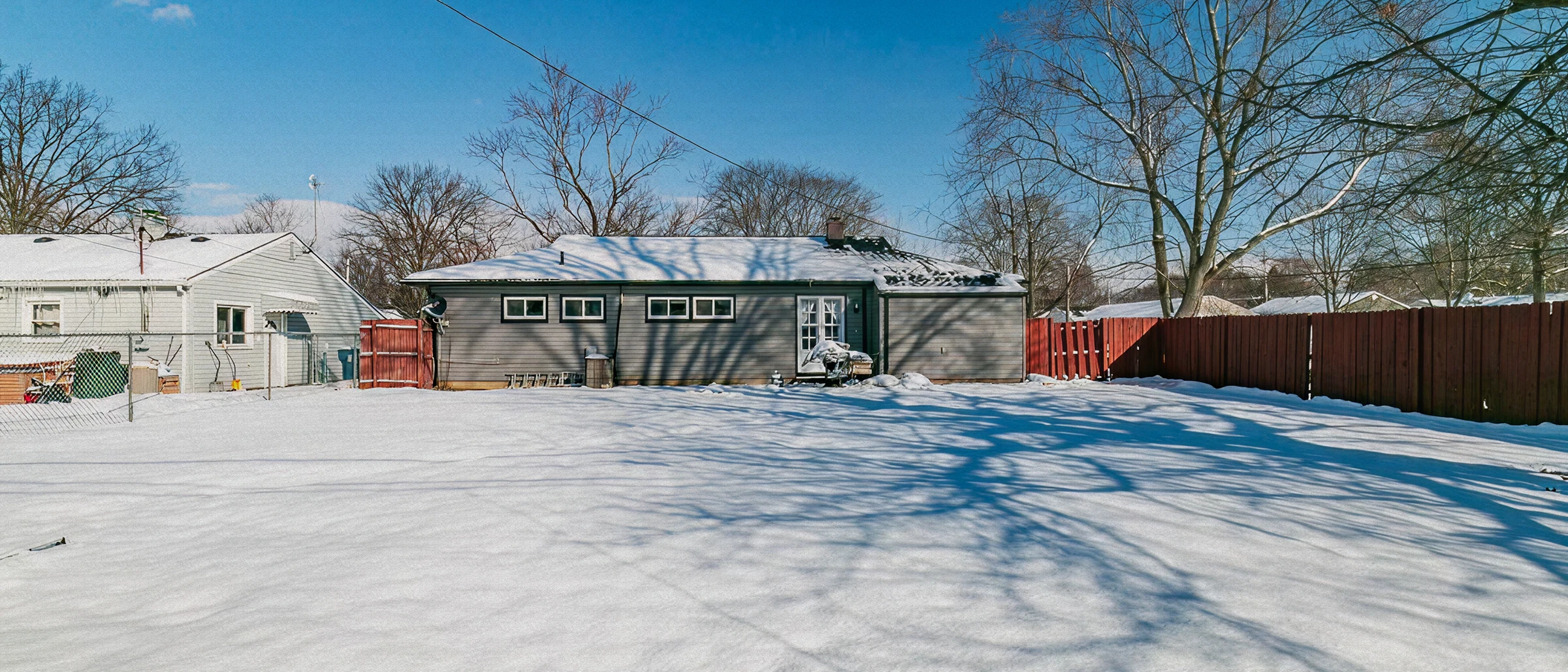 Panoramic view of a snow-covered backyard behind a single-story home with blue sky and bare trees